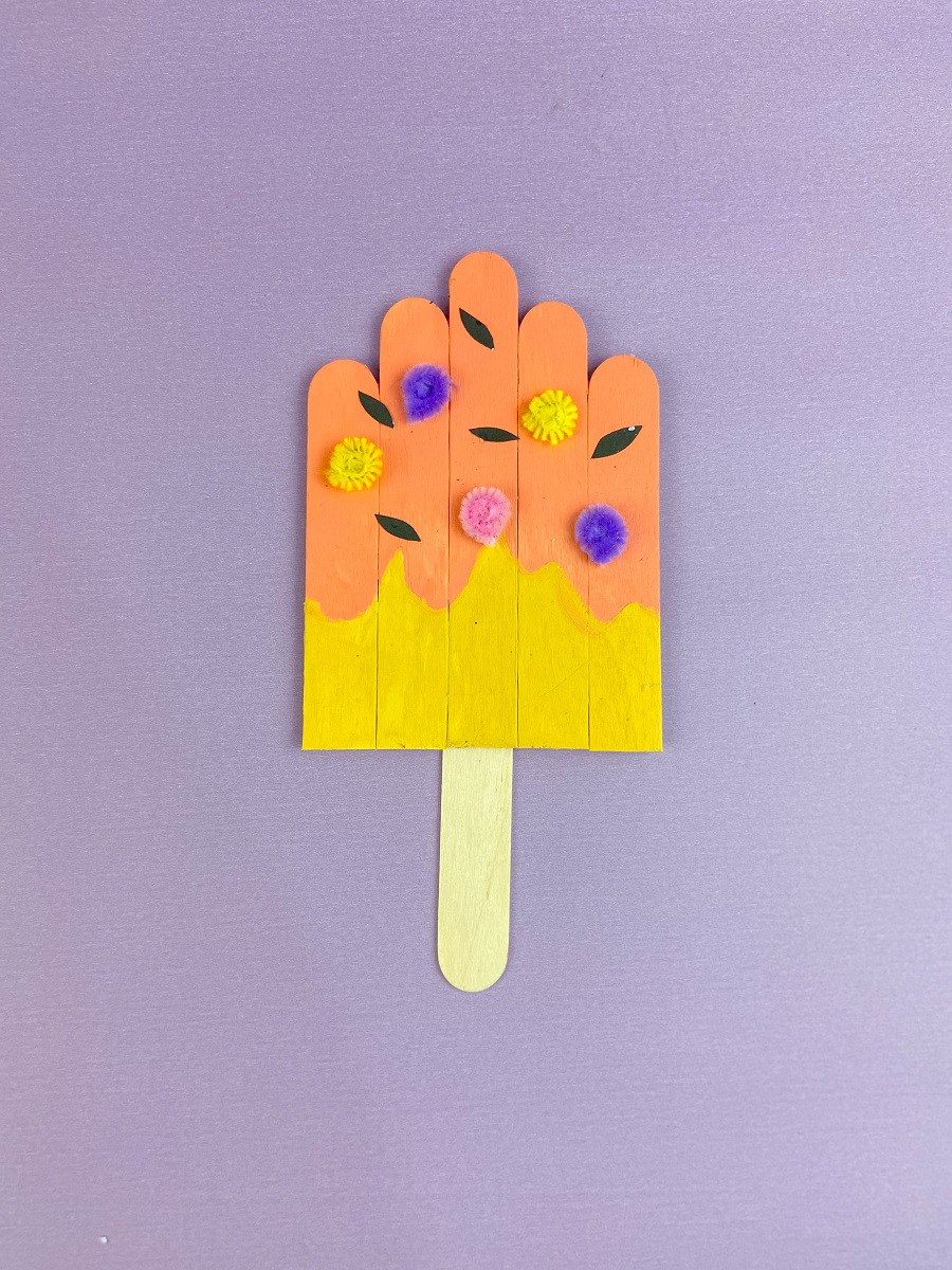 Painted craft sticks glued together to create a popsicle craft. Popsicle is decorated with pieces of paper and pipe cleaners. Completed projects lays on a purple background.