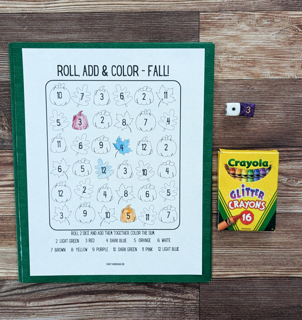 Roll, add, and color fall themed dice activity page with a couple items colored in. A box of crayons and a pair of dice lay next to the paper.