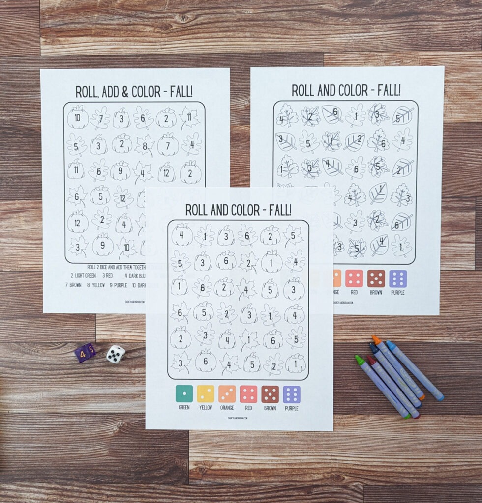 Overhead view of the fall themed roll and color math worksheets printed out. Crayons and dice lay next to the three pages.