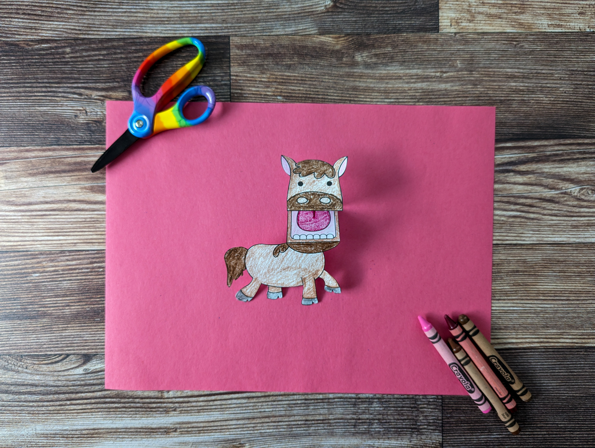 Completed horse finger puppet from set of printable farm animals. Horse is laying on a piece of red paper with scissors and crayons around it.