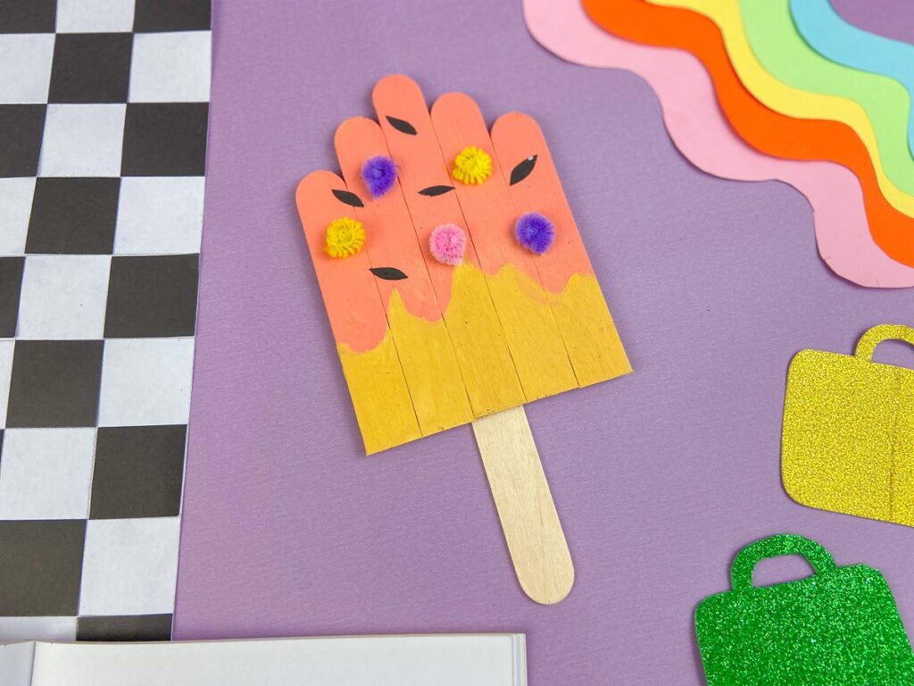 Completed popsicle craft laying on a purple background and surrounded by assorted craft supplies and back to school decorations.