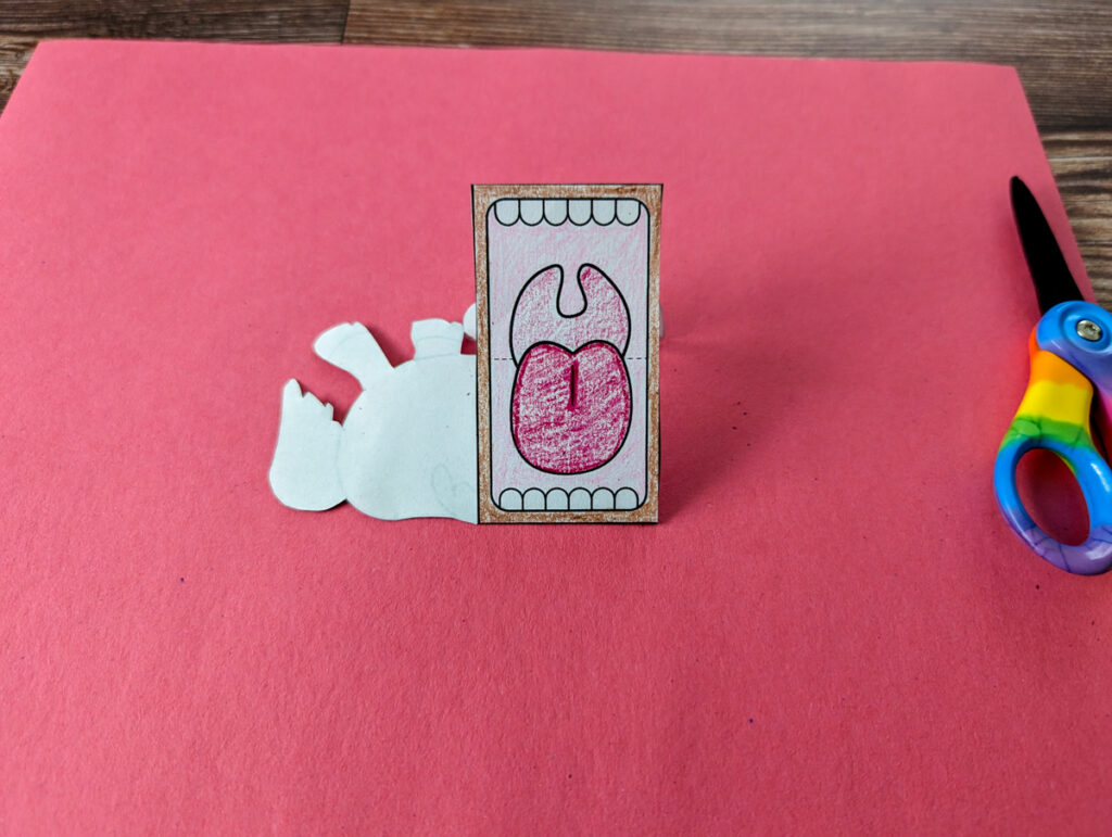 Horse colored with crayon, cut out, and mouth in process of being folded. Partially folded finger puppet is laying on a piece of red construction paper next to scissors.