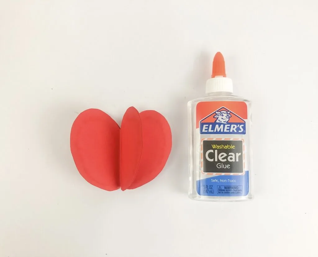 Three red pieces of paper folded and glued together to fan out in an apple shape laying next to a bottle of glue.