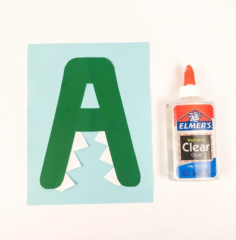 Green uppercase letter A laying on a light blue piece of paper. White teeth have been glued to the letter to look like an alligator's open mouth. A bottle of glue is on the right side.