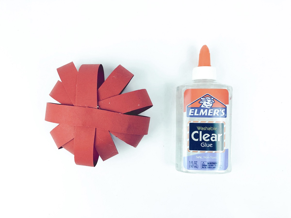 Red strips of paper glued together into overlapping circles in a ball shape. A bottle of glue lays next to it.