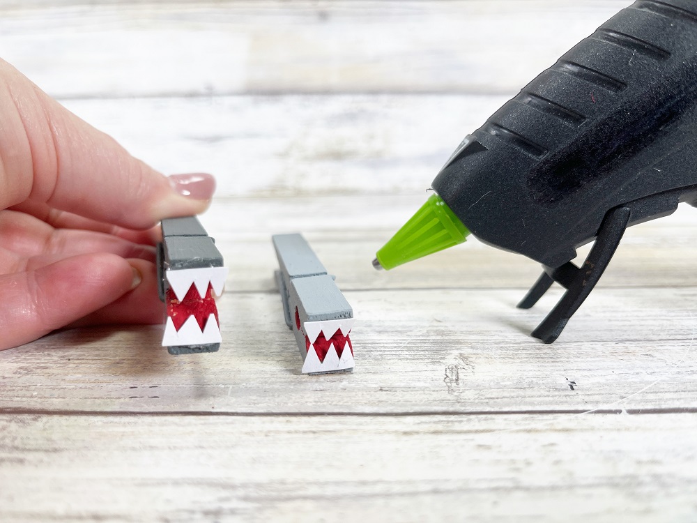 Two clothespins that have been painted gray and red have white cardstock teeth glued on to the front. Hand it holding up the one on the left. Glue gun is to the right of the other one.