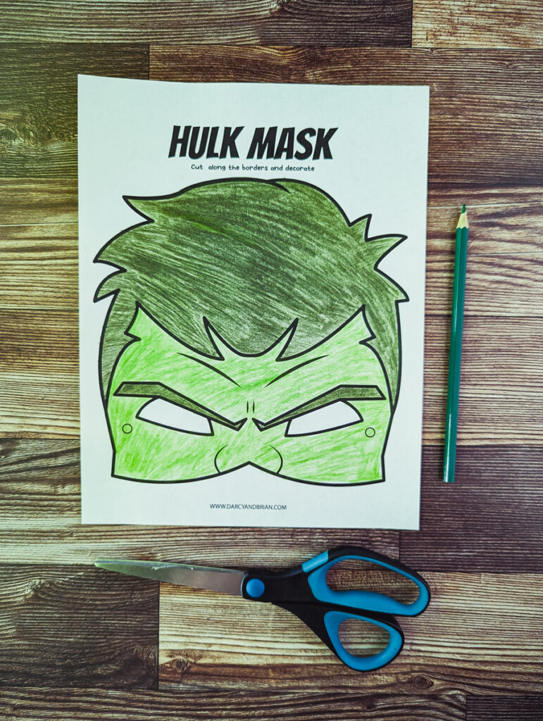 The Hulk printable mask printed out and colored in. A green colored pencil and scissors laying alongside the coloring page.