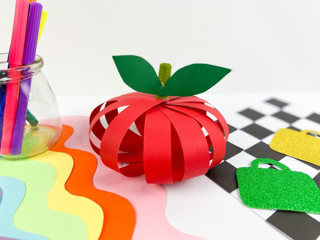 Apple made with red construction paper strips, green felt stem, and green paper leaf. It is sitting on a desk with assorted papers and markers.