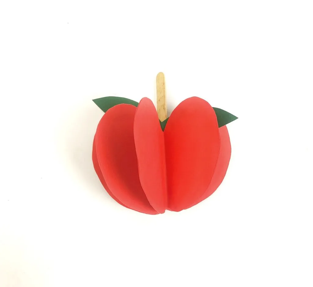 Red paper glued together to look like an apple laying against desk with popsicle stick coming out of the center.