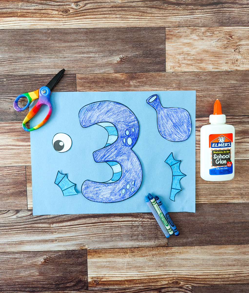 Colored in number three and other pieces to create a seahorse are cut out and laying on a piece of blue construction paper. Kids' scissors are on the upper left. A bottle of glue is laying on the right side. A few crayons are laying across the paper.