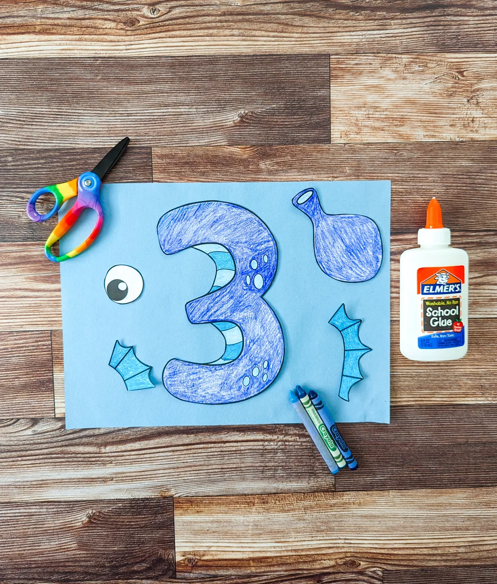 Colored in number three and other pieces to create a seahorse are cut out and laying on a piece of blue construction paper. Kids' scissors are on the upper left. A bottle of glue is laying on the right side. A few crayons are laying across the paper.