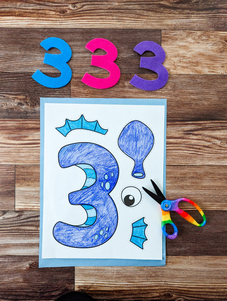 Craft template printed out and colored in. Three felt number 3s lined up above the paper. A pair of kids' scissors lays by the edge of the number 3 craft.