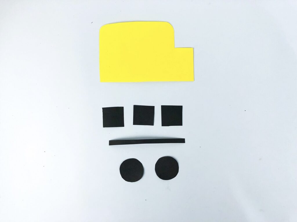 Yellow paper cut in rectangular bus shape. Black paper cut into three small squares, one thin strip and two small circles all laid out.
