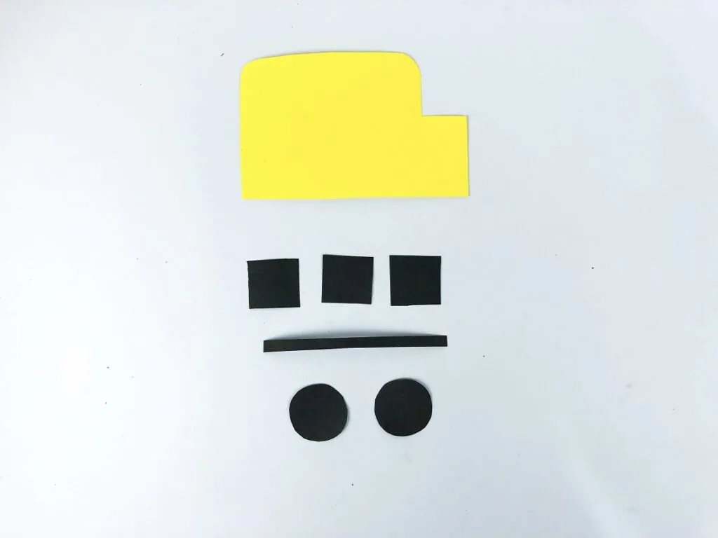 Yellow paper cut in rectangular bus shape. Black paper cut into three small squares, one thin strip and two small circles all laid out.