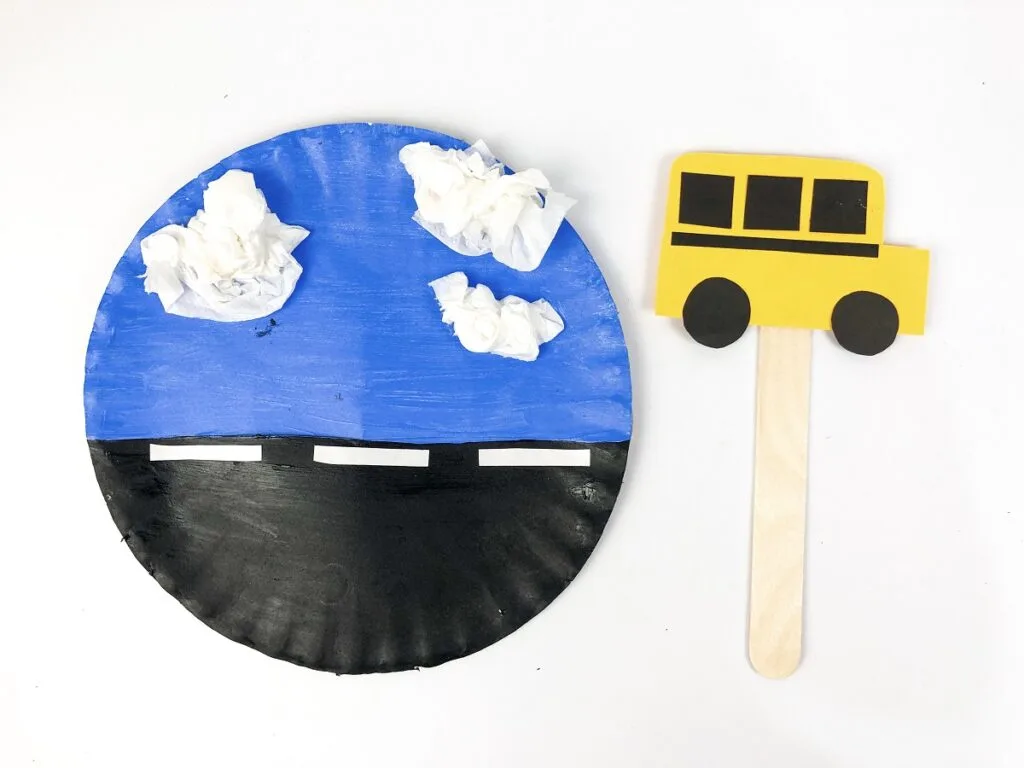 Paper plate painted half blue and half black with white tissue paper glued in puffy clumps as clouds and masking tape to make road lines. Next to plate is a small construction paper bus on a popsicle stick.