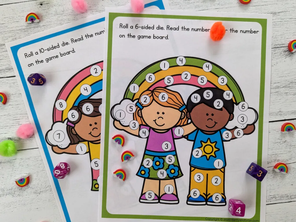 Two pages of the roll and cover rainbow game are printed out. One is for numbers 1-6 and the other is numbers 0-9 or 1-10. There are dice, mini rainbow erasers, and assorted colors of pom poms scattered around the printables.