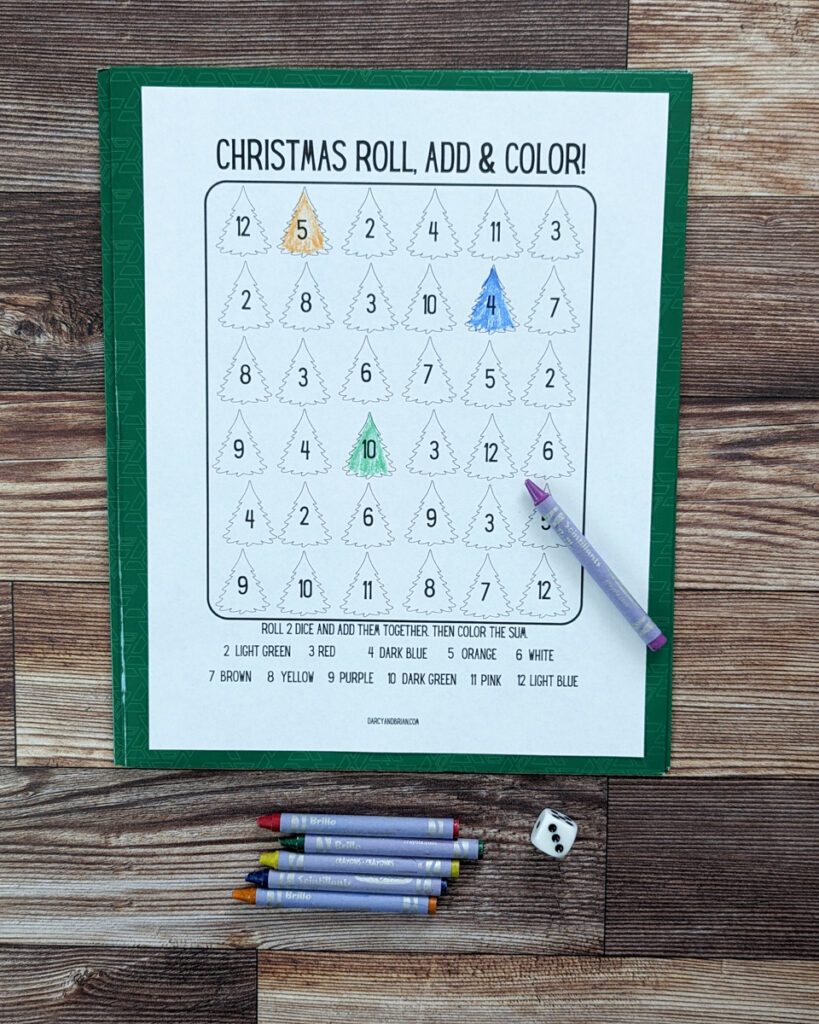 Overhead view of roll and color Christmas printable dice game addition practice page. Three different Christmas trees are colored in. Crayons laying below the game worksheet and one on it. 