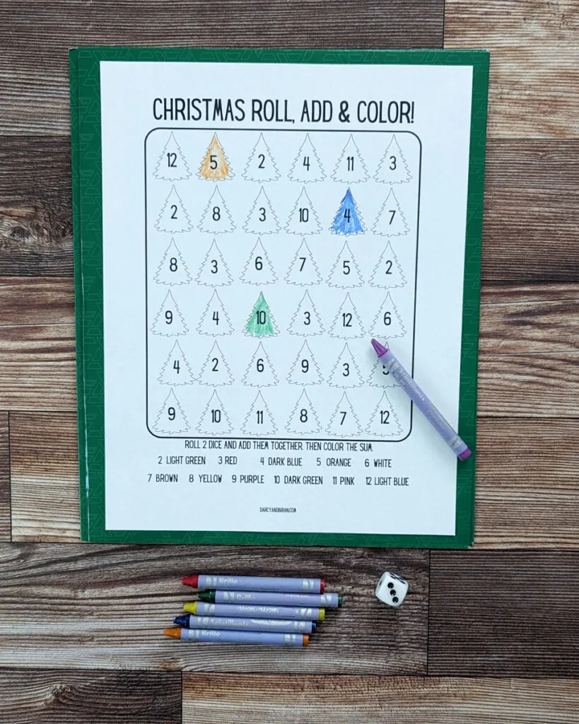 Overhead view of roll and color Christmas printable dice game addition practice page. Three different Christmas trees are colored in. Crayons laying below the game worksheet and one on it.