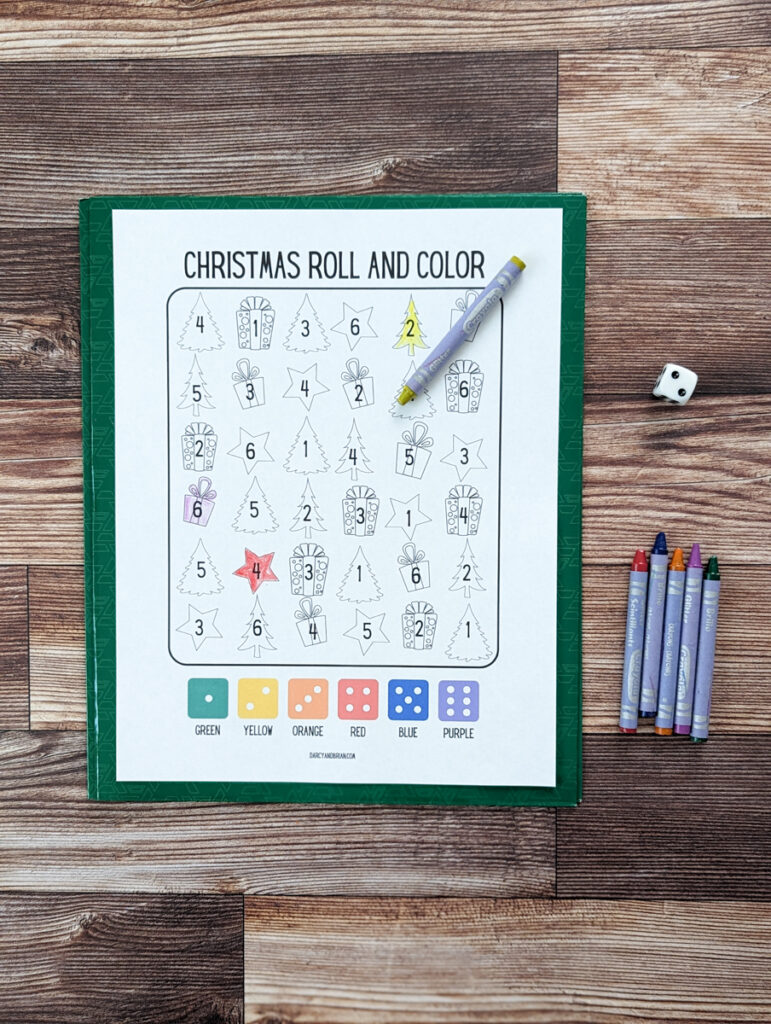 Top down view of one printed out Christmas themed roll and color page. A couple pictures are colored in. One crayon is laying on the worksheet. More crayons and a six sided die are on the right.