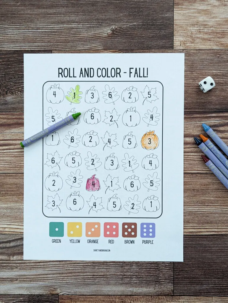Top down view of printed out roll and color fall number recognition dice game. A couple of pictures are colored in, a die is nearby, and assorted crayons.