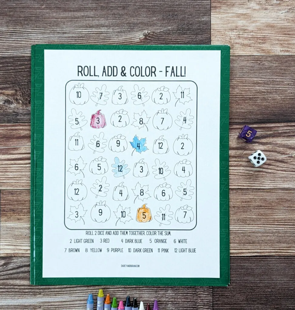Overhead view of the addition practice page of the roll and color fall printables. Several pumpkin and leaf pictures are colored in with crayon. Two six sided dice on the right side.