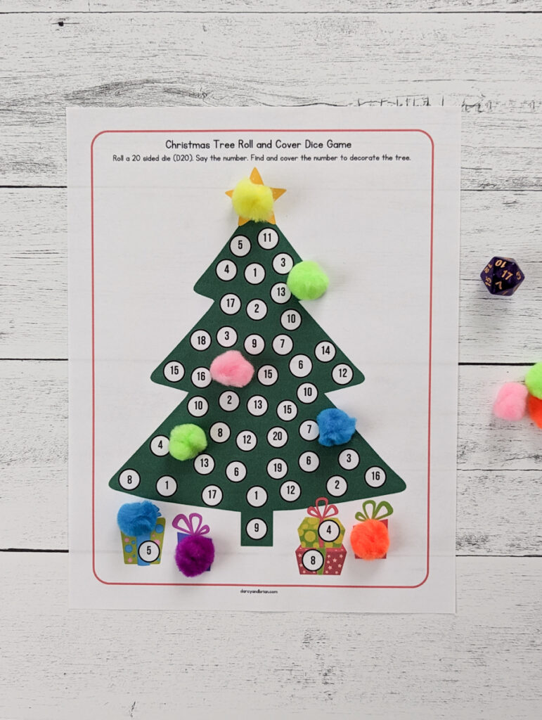 A D20 is laying next to a printed out page of the Christmas Tree Roll and Cover game. The tree and gifts have white circles with a number between one and twenty in them. Several numbers have been covered with colorful pom poms.