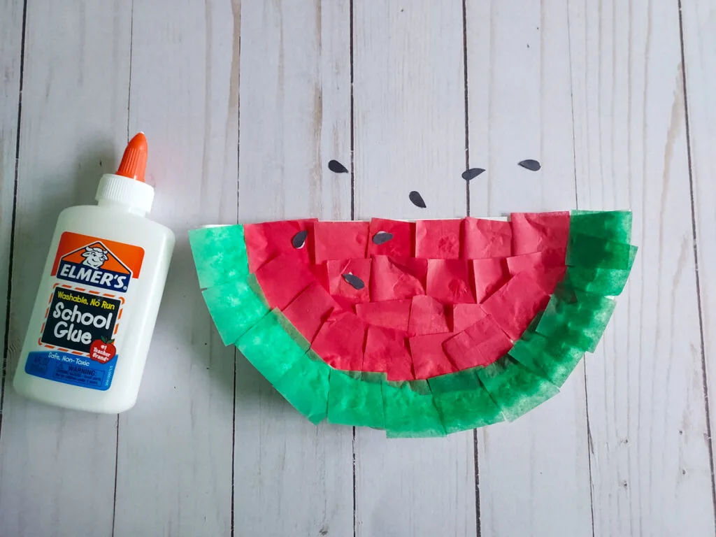 Bottle of glue laying next to half a paper plate covered in red and green tissue paper resembling a watermelon. Black pieces of paper glued on for seeds and some scattered on table.