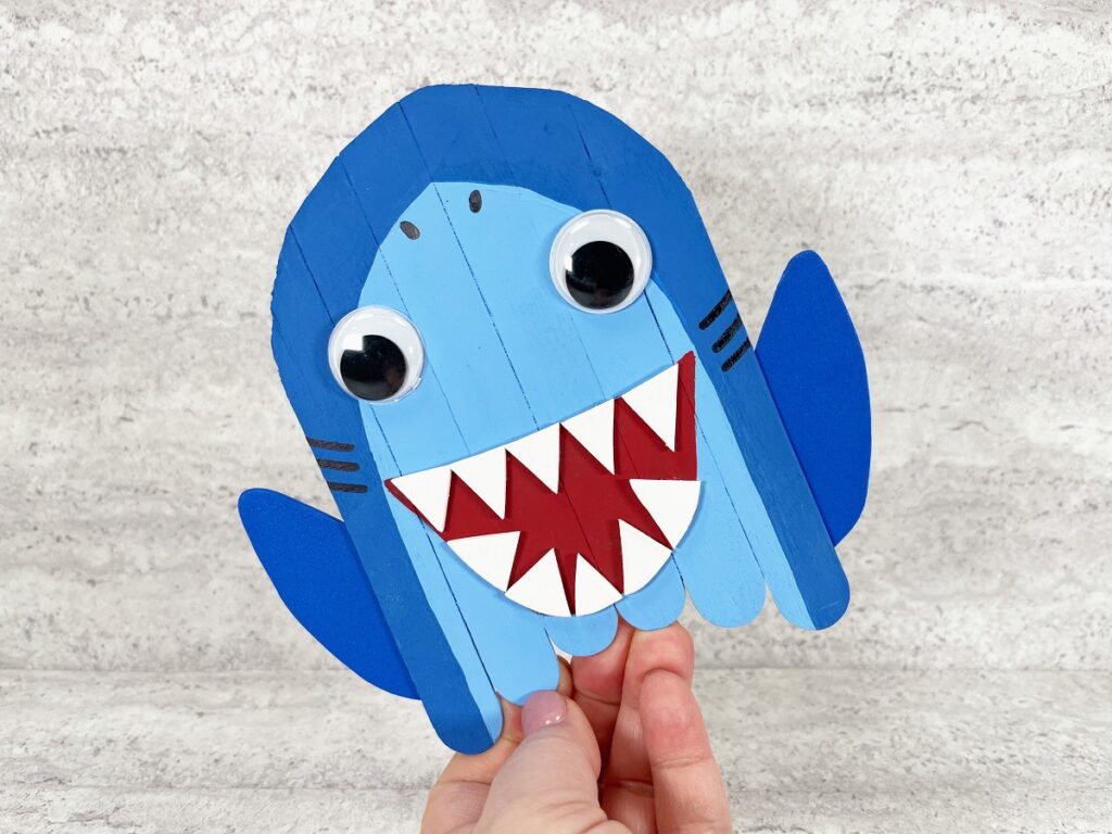 Caucasian woman's hand holding up completed popsicle stick shark craft. Shark is being held tilted toward the upper left like it is swimming.