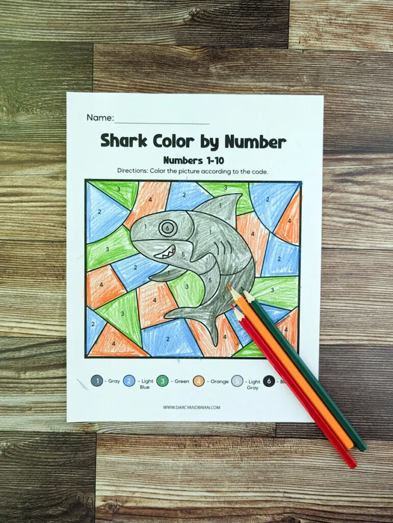 Single color by number shark worksheet printed out and colored in by a child. Three colored pencils lay diagonally over bottom right corner of the page.