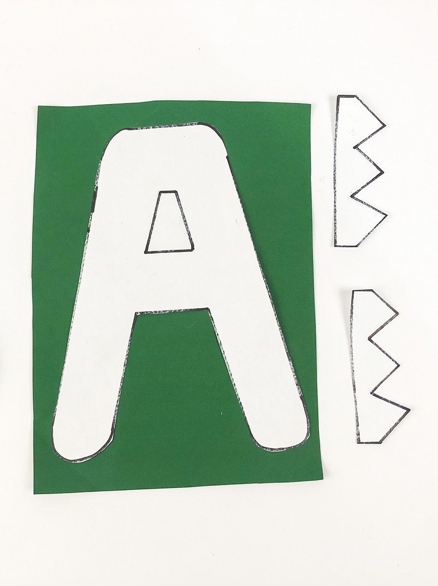 A capital letter A printed out on white paper and laying on green paper to be traced. Zig zag shapes are cut out from white paper next to it.