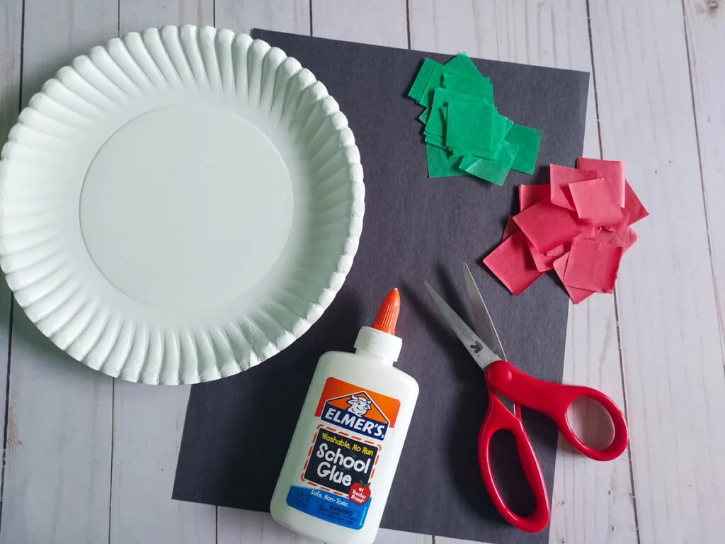 Paper plate, glue, scissors, green and red tissue paper squares, and black construction paper arranged on table.