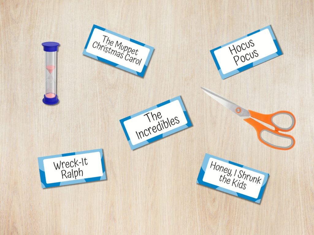 Five charade word cards with Disney movie titles spread out on a desk in a digital mockup with a pair of scissors and a small sand timer.