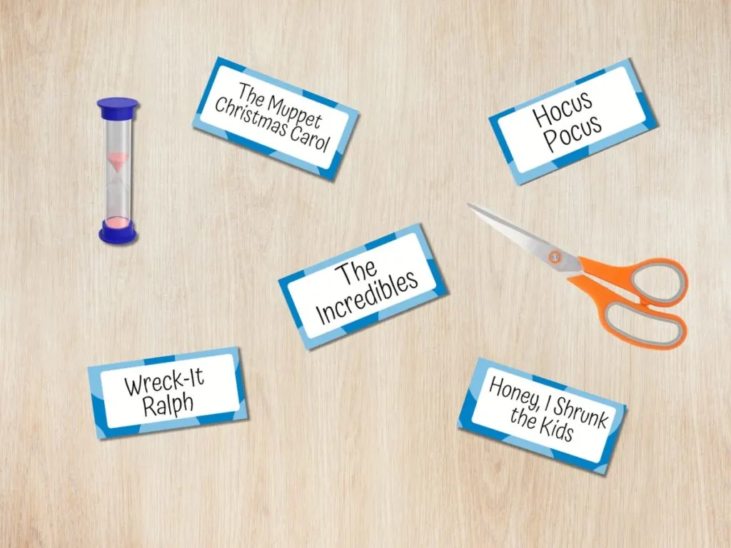 Five charade word cards with Disney movie titles spread out on a desk in a digital mockup with a pair of scissors and a small sand timer.
