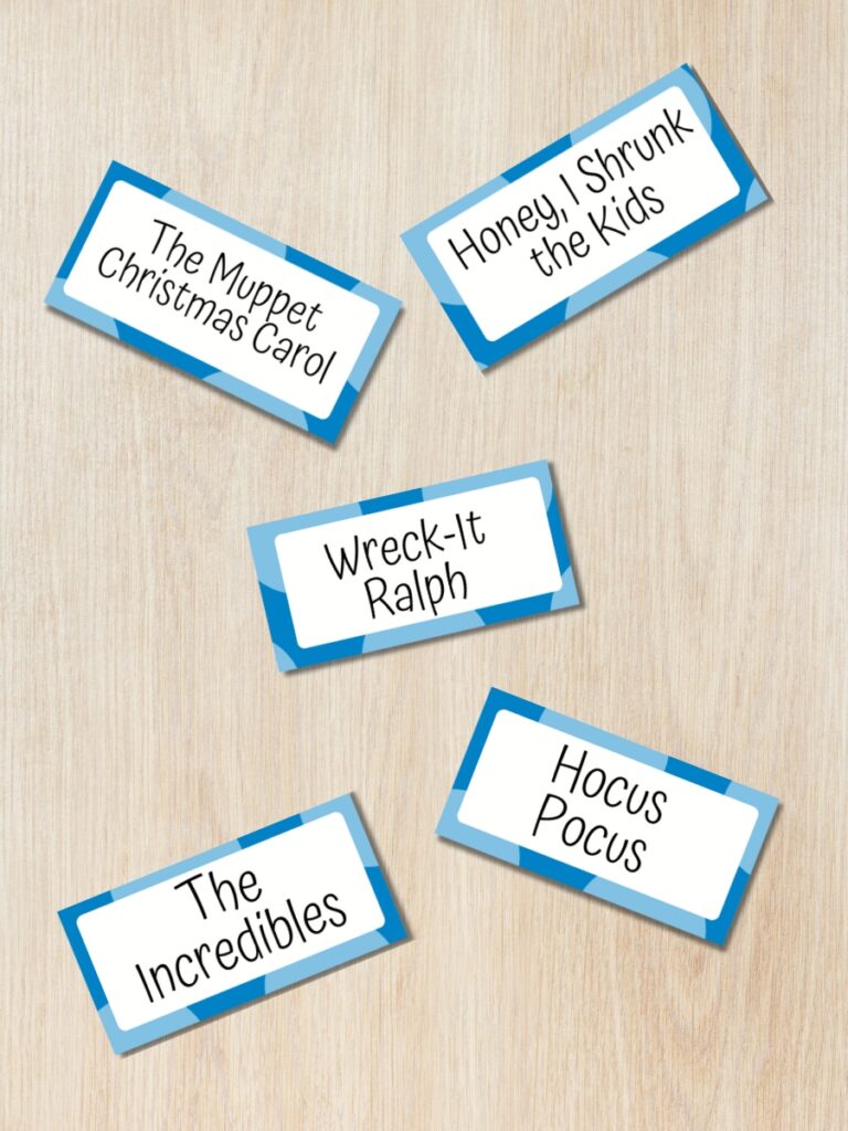 Mockup of five charades word cards spread out on a desk background. The movie titles include The Muppet Christmas Carol, Honey I Shrunk the Kids, Wreck-It Ralph, The Incredibles, and Hocus Pocus.