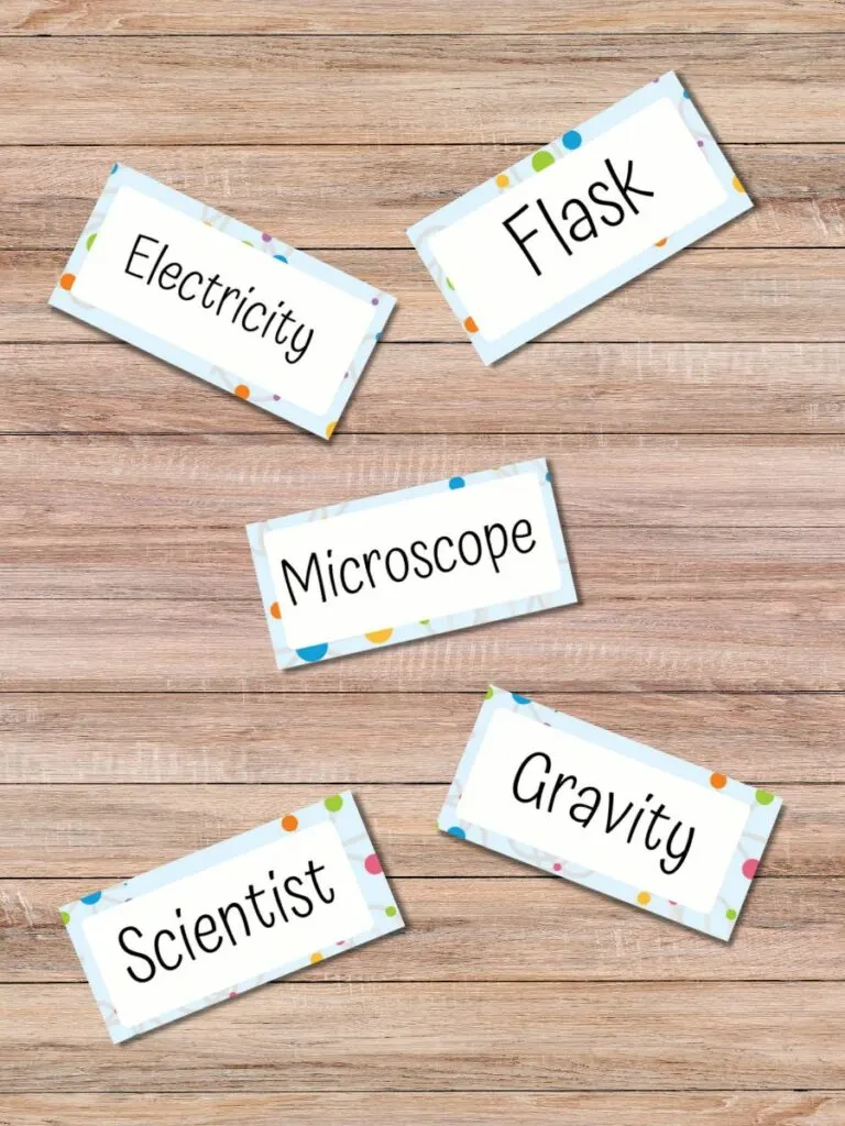 Mockup image of five Science Pictionary word cards spread out on a wooden table. The cards say Electricity, Flask, Microscope, Scientist, and Gravity.