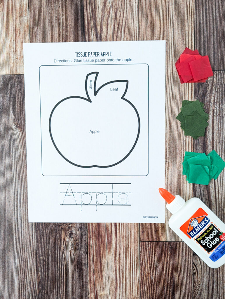 Overhead view of printed out apple craft template. Piles of pre-cut red, dark green, and green tissue paper along the right side. A bottle of glue is laying pointing towards the apple outline.