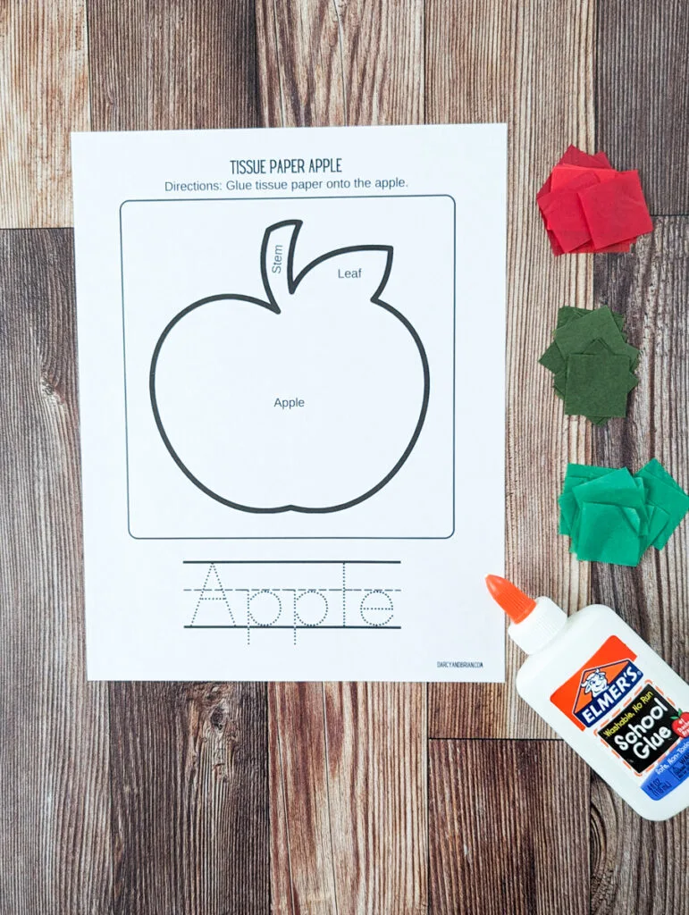 Overhead view of printed out apple craft template. Piles of pre-cut red, dark green, and green tissue paper along the right side. A bottle of glue is laying pointing towards the apple outline.