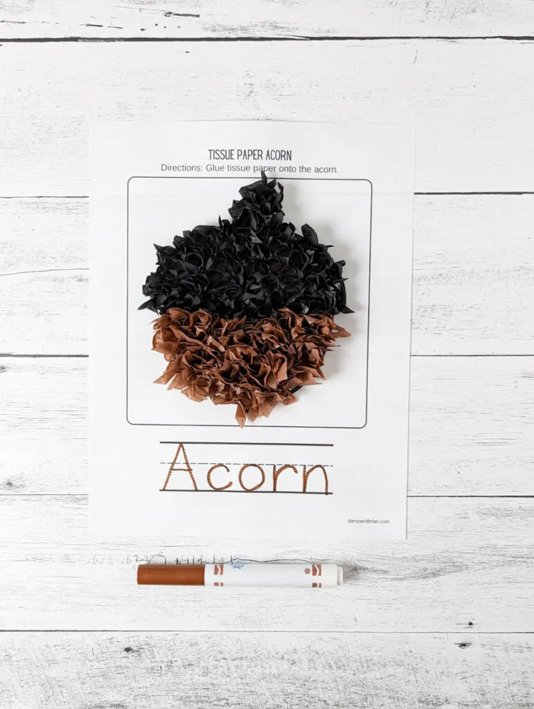 Top down view of completed acorn covered in black and brown tissue paper that is sticking up. It has a fluffy textured look. A brown marker traced the word "acorn" at the bottom of the page.
