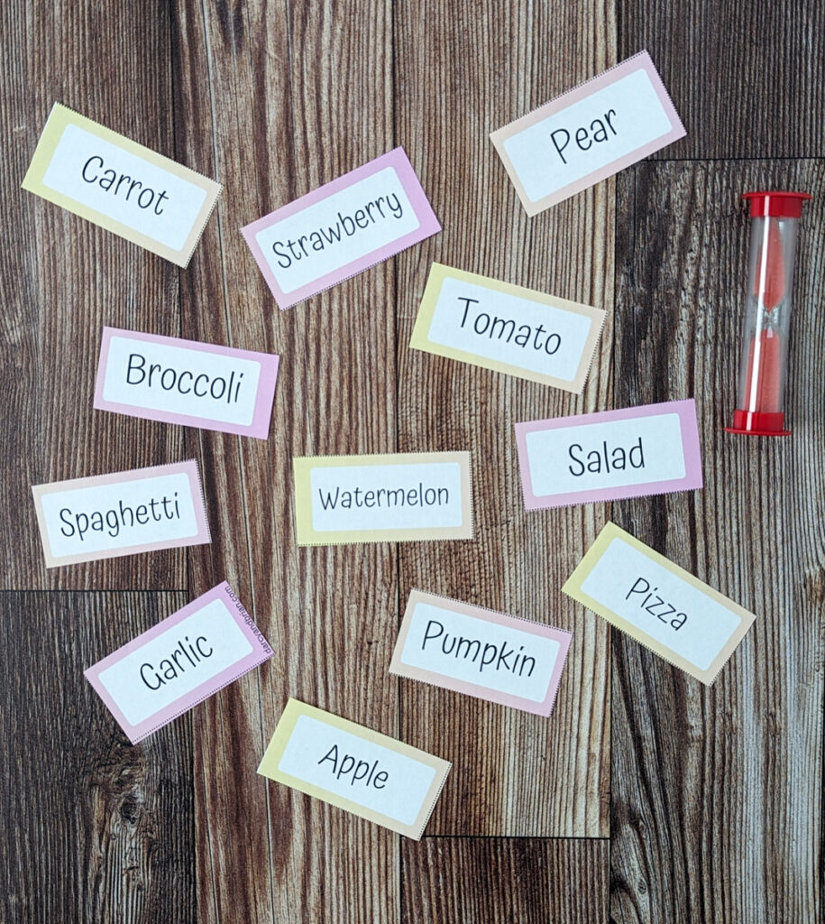 An assortment of food word cards for a Pictionary drawing and guessing game cut out and spread out on table next to red sand timer.