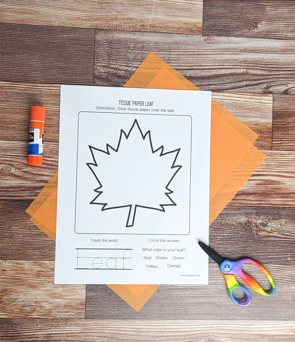 Leaf craft template printed out and laying on top of sheets of orange tissue paper. A glue stick and scissors are nearby.