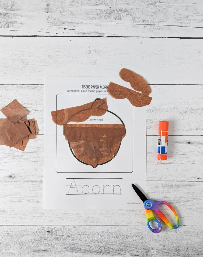 Brown pieces of tissue paper being glued down flat onto acorn template. Some strips of paper are cut with a curvy side.