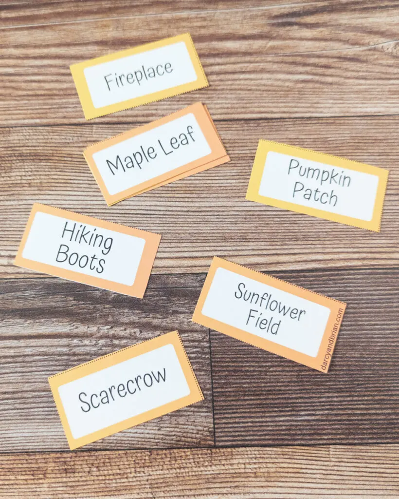 Six fall word cards for playing Pictionary lay on the table. They say fireplace, maple leaf, pumpkin patch, hiking boots, sunflower field, and scarecrow.