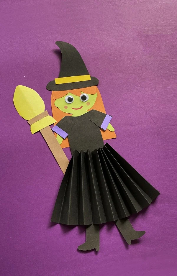 A witch holding a broom made out of paper and has googly eyes to give her a cute look.