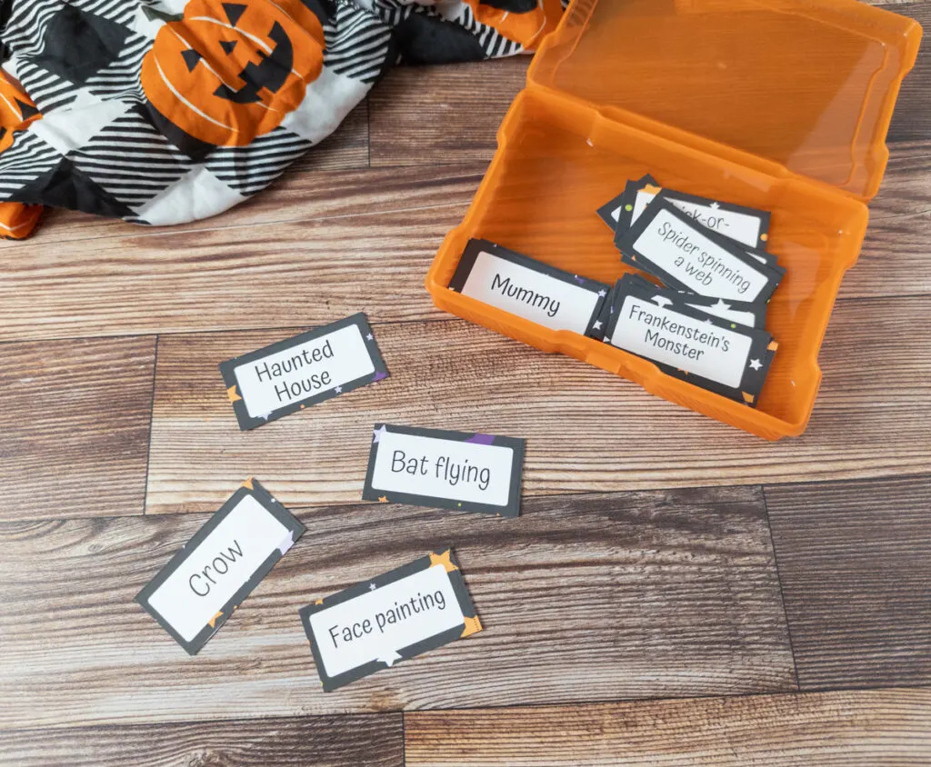 Charades cards with Halloween themed topics are cut up and in an open orange task card box. A few are laying on the table. The ones visible say: haunted house, bat flying, crow, face painting, mummy, Frankenstein's monster, and spider spinning a web.