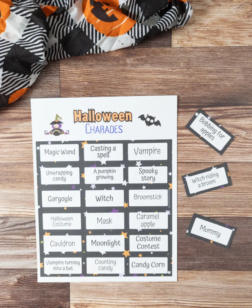 Top down view of printed out sheet of Halloween charades game prompts. A few cards are cut out and laying next to the paper. A decorative cloth with jack-o-lanterns on it at the top.