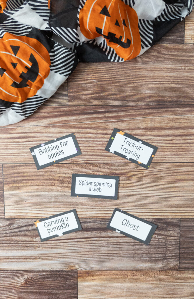 Five Halloween themed charades cards cut out and laid on table near a black and white cloth with jack-o-lanterns. Cards say: bobbing for apples, trick-or-treating, spider spinning a web, carving a pumpkin, and ghost.