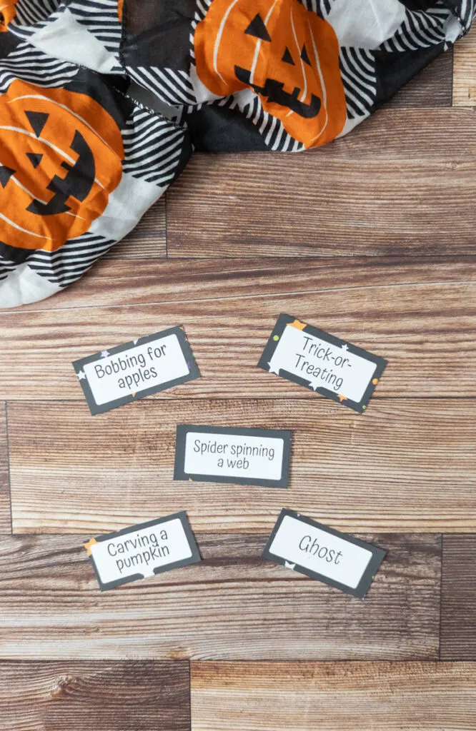 Five Halloween themed charades cards cut out and laid on table near a black and white cloth with jack-o-lanterns. Cards say: bobbing for apples, trick-or-treating, spider spinning a web, carving a pumpkin, and ghost.