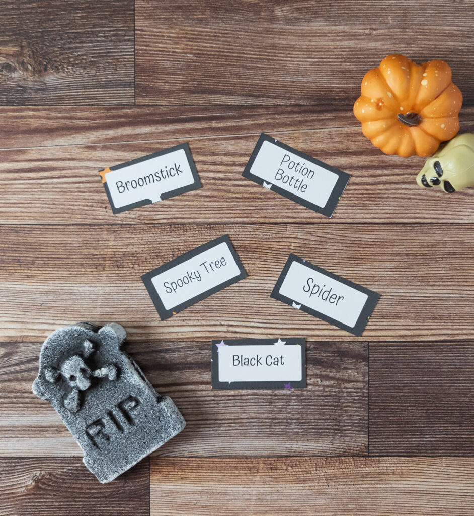 Five Halloween Pictionary cards spread out on table. Cards say: broomstick, potion bottle, spooky tree, spider, and black cat. A small pumpkin, tombstone, and skull are arranged as props.