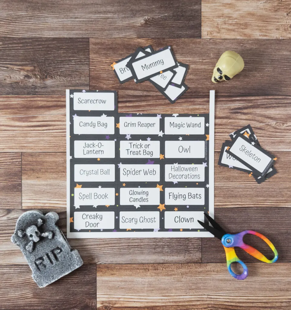 Halloween themed Pictionary drawing prompts being cut out of the printable page. A pair of scissors, a small fake tombstone, and a small plastic skull are arranged around the cards.