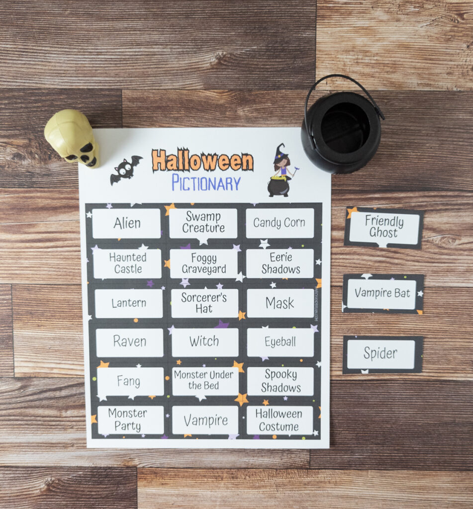 One printed out page of Halloween Pictionary prompts and three cut out cards next to it. A small black cauldron and small plastic skull are sitting on each side of the paper.